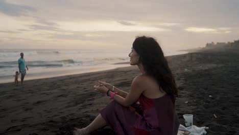 Mujer-Encantadora-Sonriente-Sentada-En-La-Arena-De-La-Playa-Con-Olas-Salpicadas