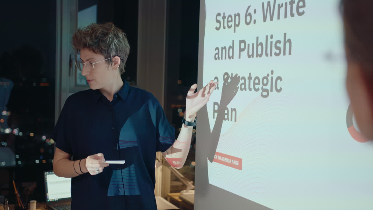 joven mujer de negocios de pie con su colega por la pantalla de proyección y dando una presentación durante una reunión en la oficina tarde en la noche. sobre el hombro disparado