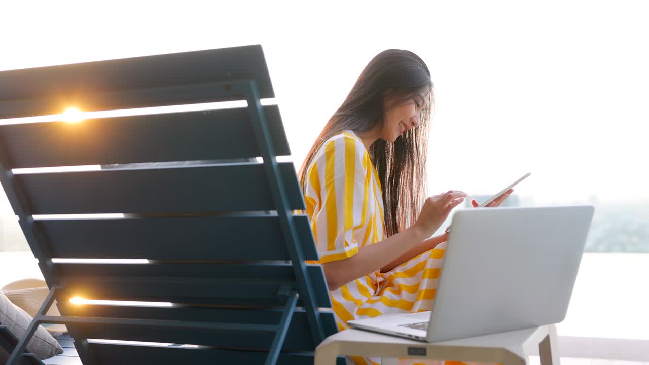 젊은 아시아 여성은 휴식 분위기에서 고객과 함께 태블릿과 노트북에서 비디오 컨퍼런스를 사용하여 일하는 아이디어를 생각하고, 스카이 스크래퍼에서 도시의 전망을보고 의자를 통해 밝은 빛을보고 있습니다.