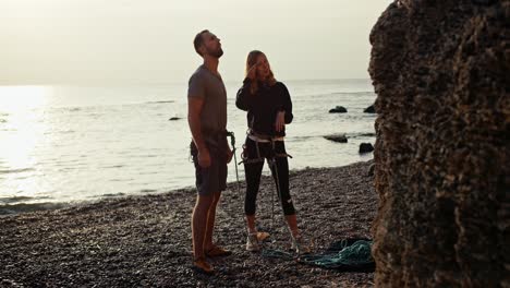 A-blonde-girl-with-curly-hair-in-a-black-Sweater-tells-a-guy-in-a-gray-T-shirt-how-they-will-climb-a-rock-near-a-rocky-seashore-and-then-insures-the-guy-so-that-he-climbs-up