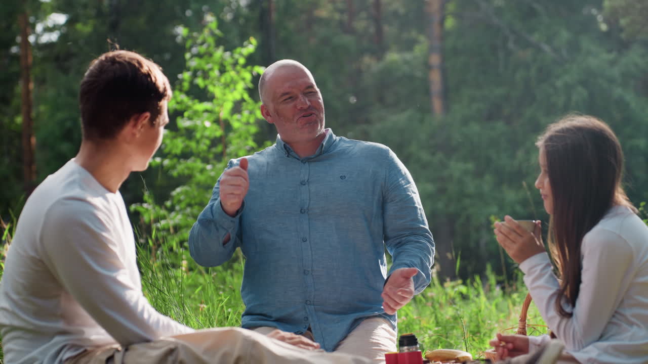 Happy father enjoying outdoor picnic with son and daughter, smiling while discussing warmly, girl sipping tea from cup and boy nodding with cheerful expression under sunlight
