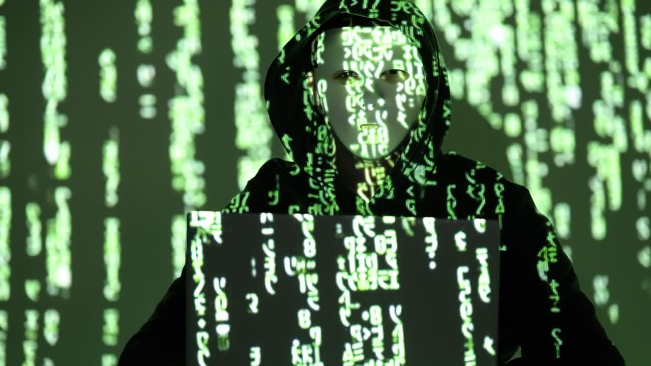 A cybercriminal in a dark hoodie and white mask stares ahead behind a laptop, illuminated by cascading green code. The scene reflects anonymity, cybercrime, and digital data infiltration