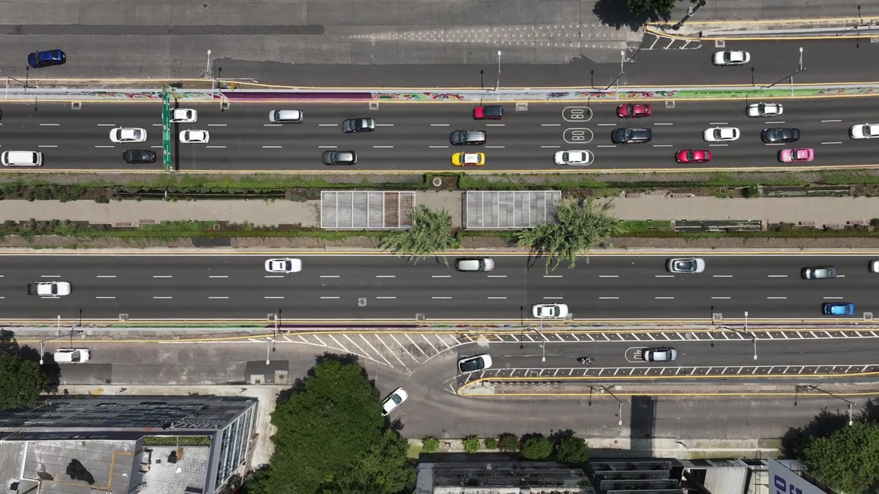 Vista aerea dall'alto con drone in ascesa verticale su un trafficato viale cittadino a più corsie con traffico scorrevole, uno spartitraffico verde e segnaletica orizzontale