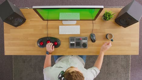 vista aérea de un editor de video masculino con auriculares inalámbricos y trabajando en una computadora con pantalla verde en una oficina creativa bebiendo una taza de café - filmado en cámara lenta