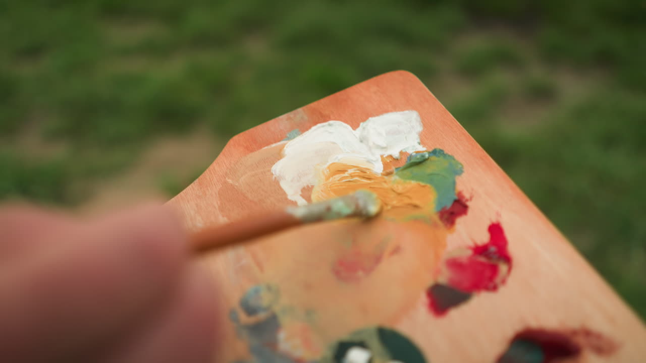 A close-up shot of an artist's hand holding a palette and mixing paint with a small brush. The vibrant colors blend together as the artist prepares to create