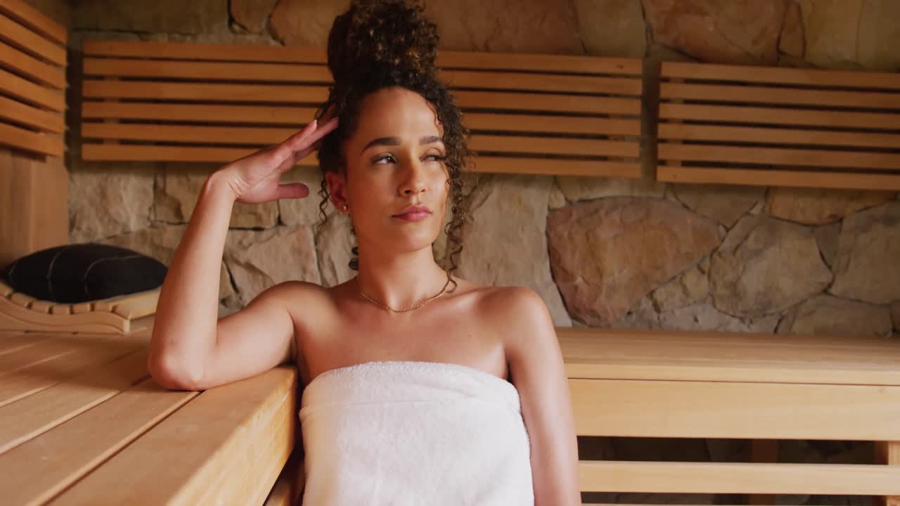 Video di una donna birazziale rilassata che indossa un asciugamano seduta in una sauna in una spa.