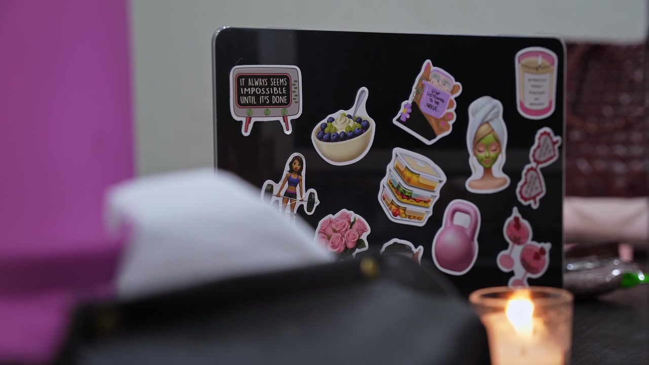 Scatto di un laptop nero coperto di adesivi a tema benessere e fitness appoggiato su una scrivania all'interno. Una candela accesa sfocata e oggetti creano una profondità di campo ridotta in un accogliente ambiente di lavoro domestico