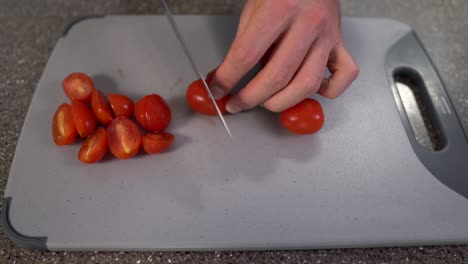 man hands cutting the cherry tomatoes into half pieces with chefs knife on grey chopping board on the table, top view