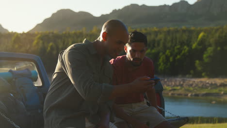 two male friends with backpacks in pick up truck by lake drinking beer watching sports on phone