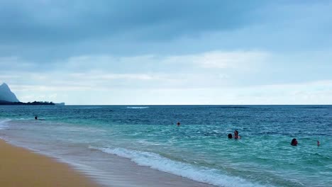 hd hawaii kauai 슬로우 모션 와이드 샷 팬은 왼쪽 하단의 해변에서 왼쪽에서 오른쪽으로 바다에 있는 몇 사람을 지나 부분적으로 흐린 하늘이 있는 아래쪽의 빈 바다로 이동합니다.