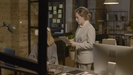 woman emplooyee at desk in the office wearing virtual reality glasses while her coworker is controlling her with tablet 1