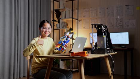 smiling teen asian girl with a cyborg hand showing thumbs up gesture to the camera while working on a laptop at home