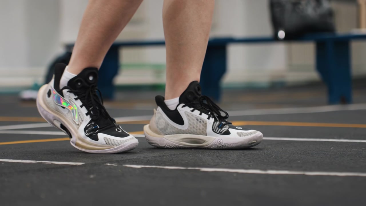 Ejercicio técnico de juego de piernas de bádminton, jugador hábil demostrando pasos controlados de talón a punta, detalle de zapatilla, ritmo tranquilo, perfil de ángulo bajo, fondo de banco, énfasis en el equilibrio y la precisión