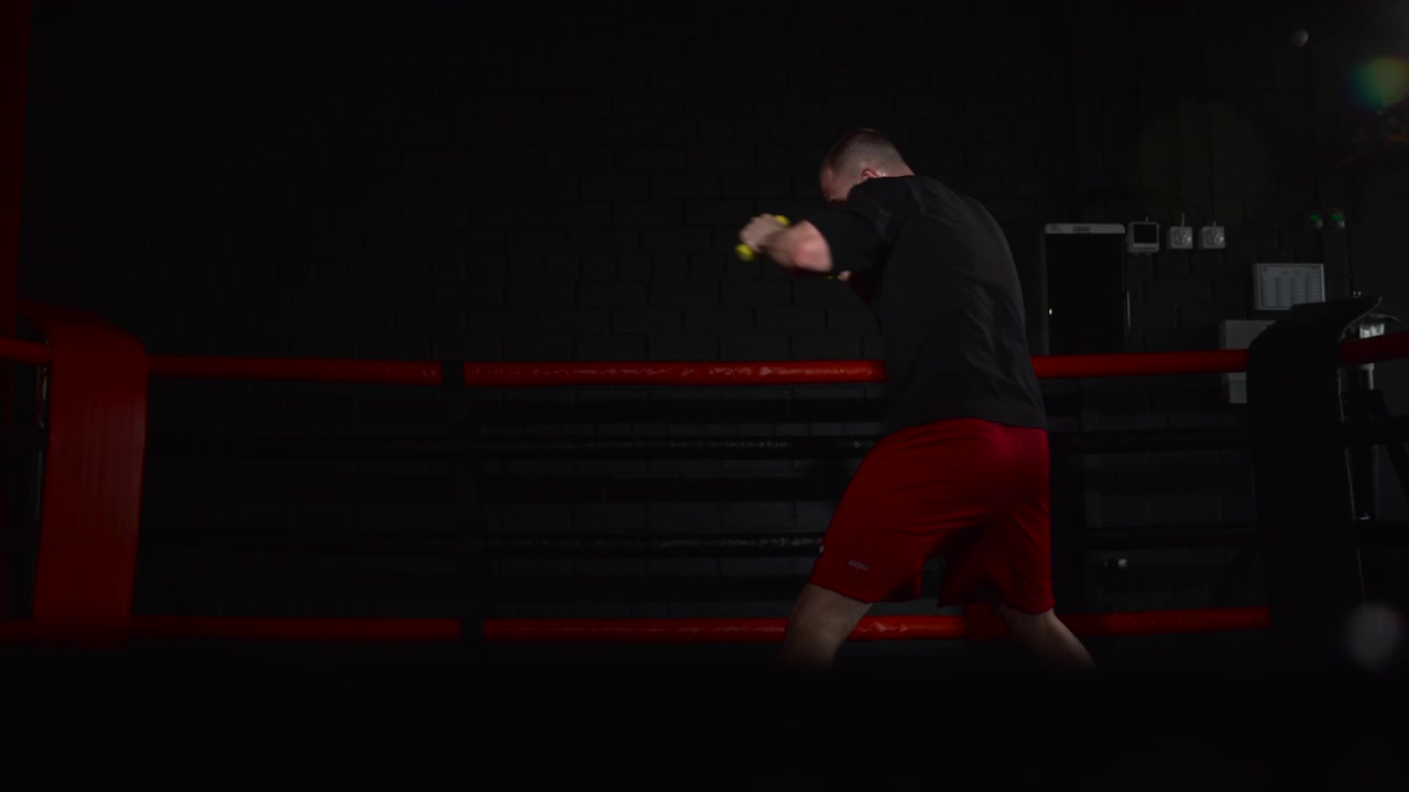 Vista posteriore a tre quarti di un pugile maschio adulto che sistema le fasce gialle per le mani mentre si appoggia alla corda superiore all'interno di una buia palestra di boxe, con dettagli rossi del ring, ombre marcate, concentrazione intensa e un'atmosfera sportiva commerciale