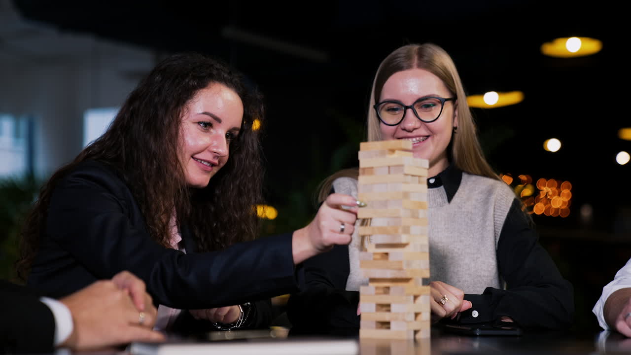 Long-haired beautiful young woman pulls a wooden brick from a pile. Blonde lady sitting next watches her attentively smiling.