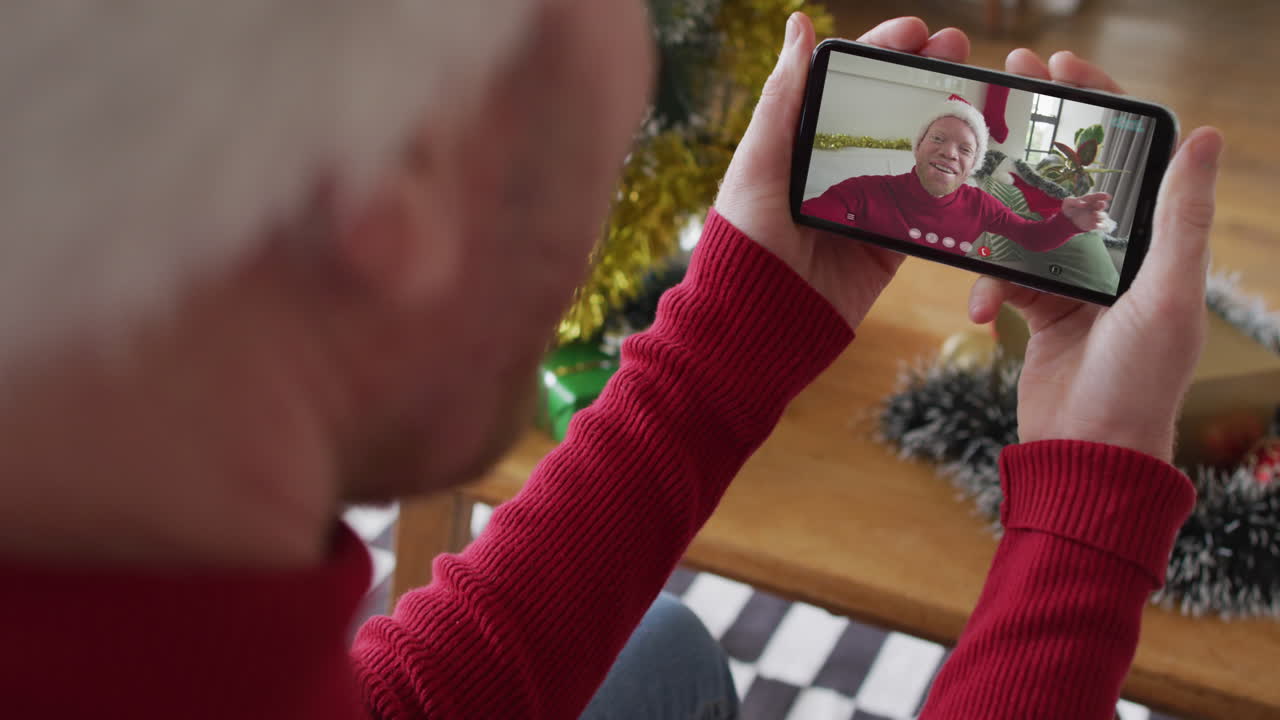 hombre caucásico con sombrero de santa usando un teléfono inteligente para una videollamada de navidad con un hombre sonriente en la pantalla