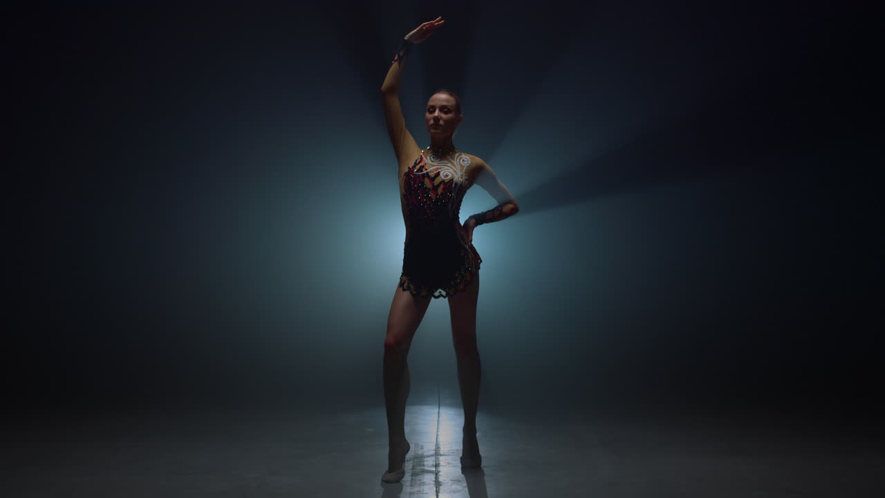 una gimnasta de pie con la mano levantada en un espacio oscuro. una deportista posando en el interior.