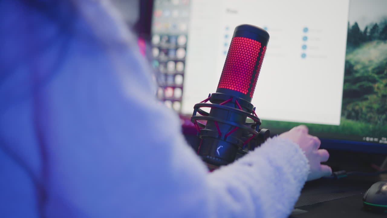 close up rear view of lady in fluffy jacket reaching for red condenser microphone on stand beside rgb light strip speaker and keyboard adjust usb cable before streaming