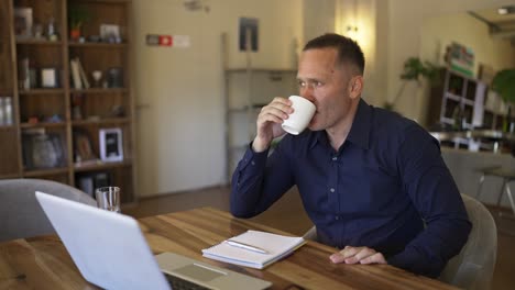 y el hombre adulto está sentado en una computadora portátil. y el hombre adulto toma un sorbo de café y se apoya en su silla