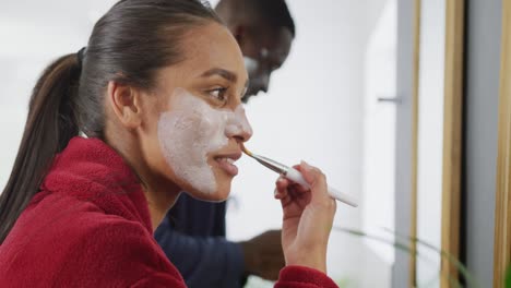 Video-of-happy-diverse-couple-applying-face-cream-and-cleansing-face-mask-at-mirror-in-bathroom