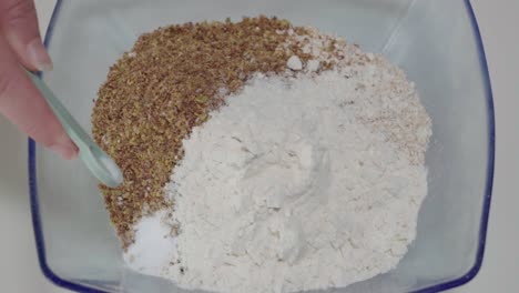 adding teaspoon of baking soda to flour and flax mixture in glass bowl, overhead