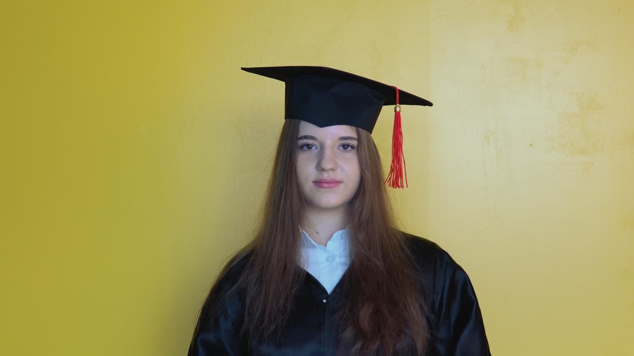 Caucasian girl student corrects the hat on her head while staying in front of camera with graduating diploma in her hands. Master of Education