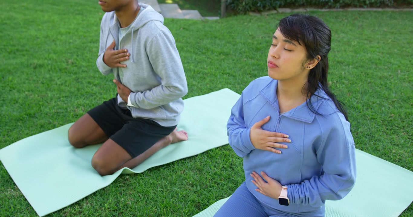 Dua orang beragam berlatih pernapasan di atas matras rumput, tangan di dada, wanita memeriksa jam tangan pintar.