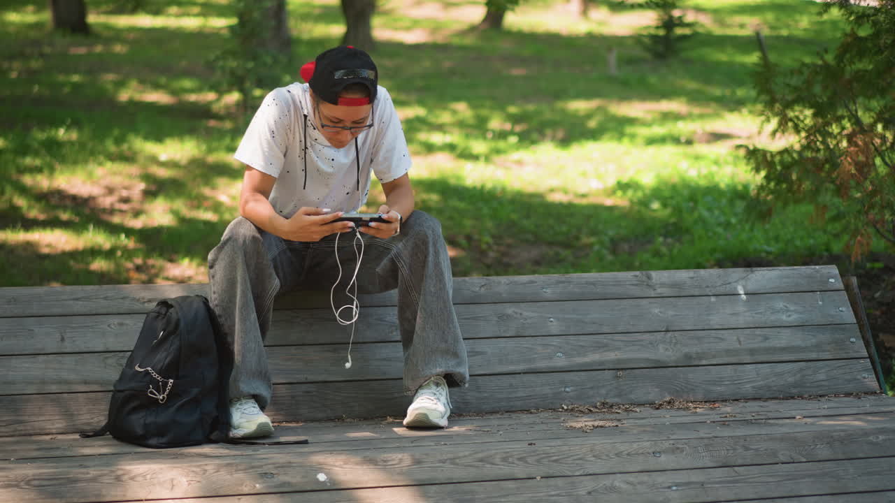 나무 벤치에 앉아 젖은 휴대폰을 확인하는 백인 남성, 물이 튀어 폰이 흠뻑 젖은 후 걱정스러운 표정으로 가방과 모자를 쓴 젊은 도시 통근자, 그늘진 나무와 공원 캐노피가 차분한 색조를 만들어 냅니다.