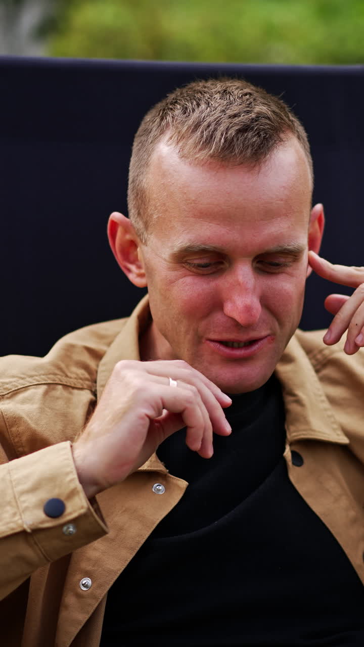 Positive relaxed man looks at the screen of his laptop. Male freelancer sitting comfortably in a garden chair working remotely from nature. Vertical video