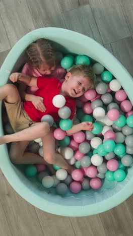 friendly little girl and boy siblings have fun lying in dry pool with plastic balls. blonde sister hugs younger brother laughing with satisfaction