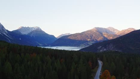 aerial-over-a-small-street-through-a-forest-in-a-valley-in-the-morning-with-dramatic-mountains-in-the-background