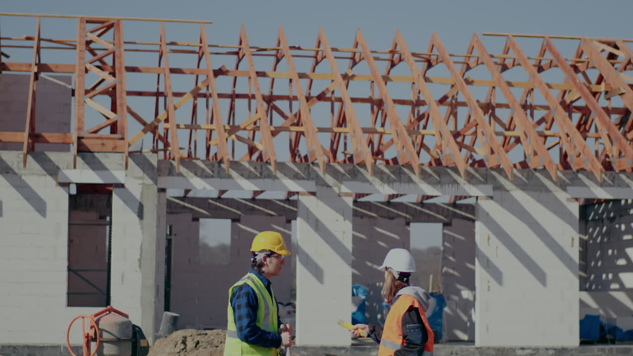 Side view of young female engineer pointing at incomplete roof beam discussing with male coworker at construction site on sunny day