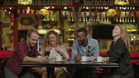 Ein-Schöner-Hispanischer-Mann-Zeigt-Seinen-Freunden-Interessante-Sachen-Auf-Seinem-Smartphone,-Während-Sie-Sich-In-Der-Bar-Amüsieren.-Sie-Lachen,-Scherzen-Und-Trinken-In-Einer-Stylischen-Hipster-Bar.