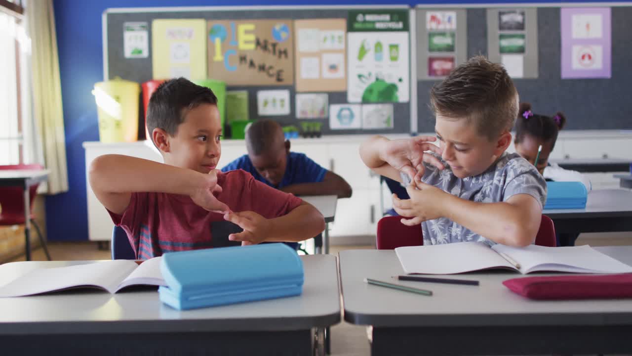 dos escolares diversos sonriendo mientras están sentados en el aula jugando con las manos