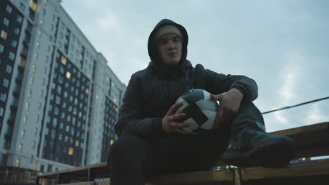 young man in hoodie sitting on bench gripping soccer ball with intense focus, rotating it during urban training session, surrounded by modern residential buildings and evening lights