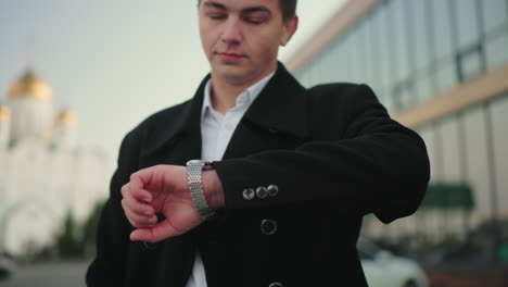 investor in black coat checking time with confident look and right hand in pocket, standing in front of modern glass building, successful businessman exuding confidence in urban setting