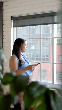 vertical video: holding tablet, businesswoman in blue blouse standing by window, looking outside