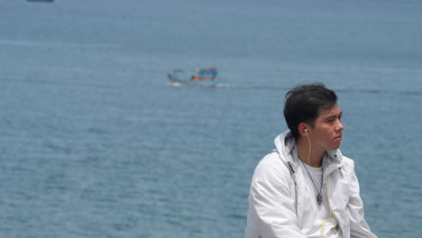 Un-Joven-Modelo-Asiático-Escucha-Música-Con-Un-Barco-Sellando-El-Mar-En-El-Fondo,-Estilo-De-Vida-Retrato-En-Cámara-Lenta