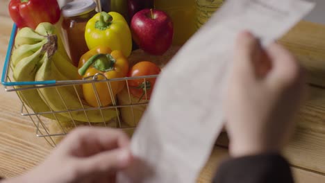 person checking bill or receipt for supermarket shopping basket full of basic food items