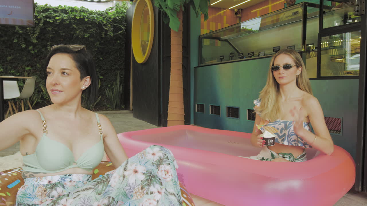 Two young women enjoy ice cream while relaxing in an inflatable pool in front of a modern ice cream parlor with tropical decor.