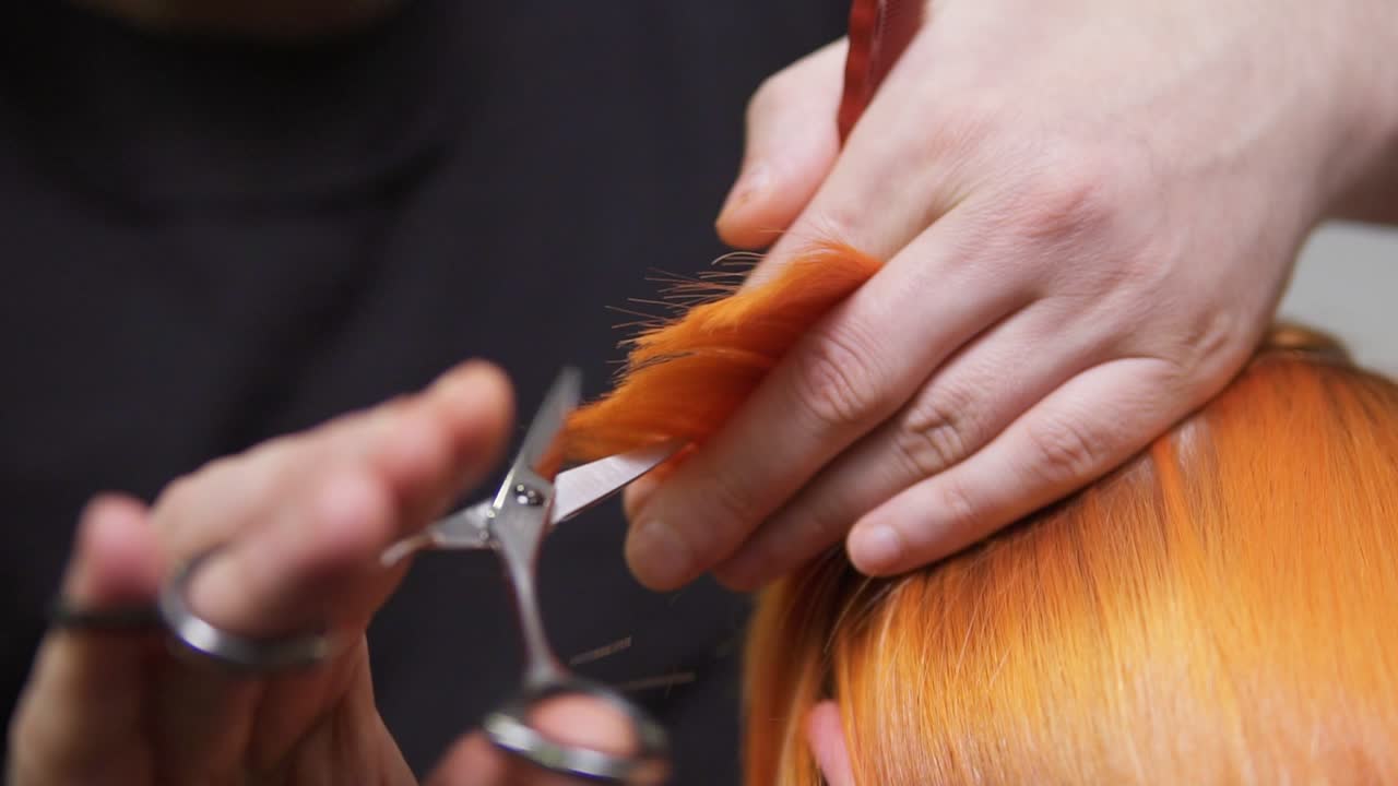 Close Up view of redhead woman's hair being cut by a professional hairdresser in beauty salon. Male hands holding a hair strand