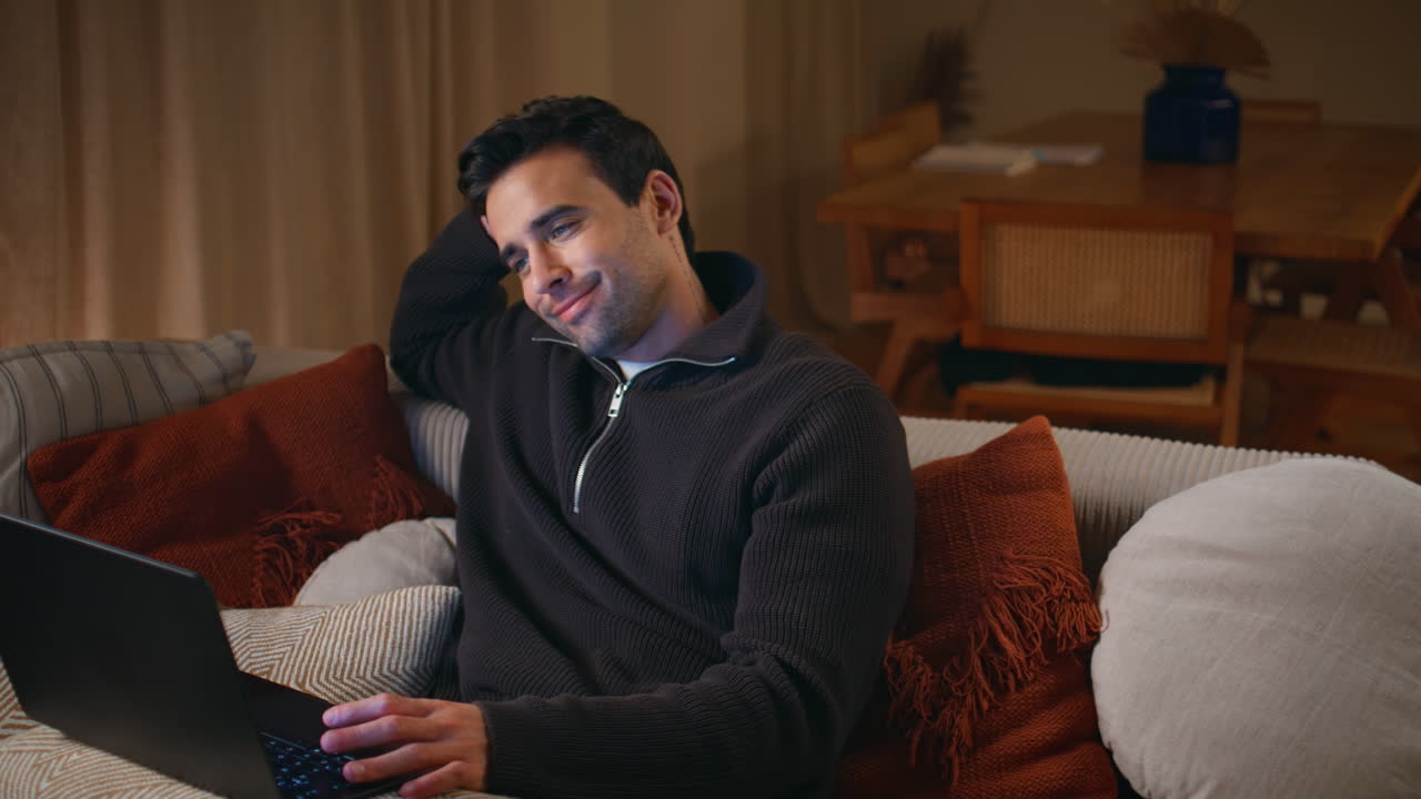 Joyful guy calling laptop on comfortable sofa at evening. Cheerful man talking