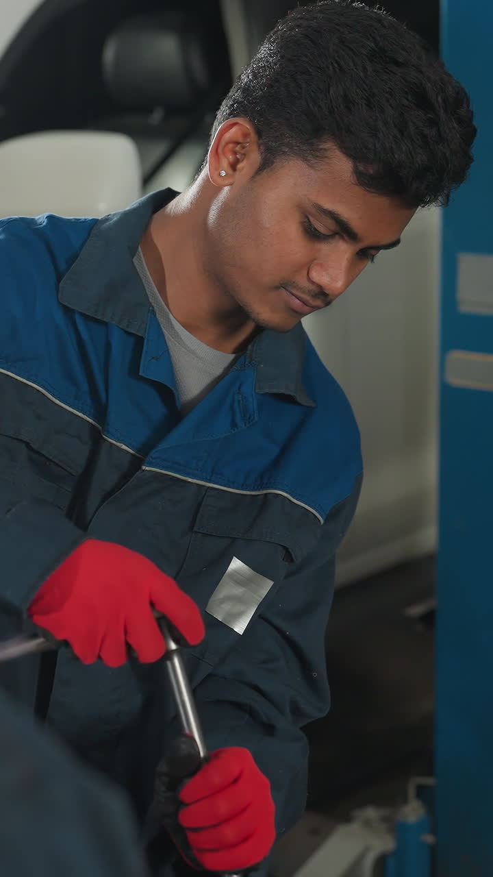 mecánico en uniforme azul y guantes rojos apretando pernos en el motor del coche mientras el colega observa desde atrás, el fondo presenta a alguien pasando en negro cerca de un ascensor de vehículo en un taller automotriz