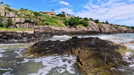 Waves-splashing-against-the-rocks,-showing-mansions-on-top-of-the-hill-in-Punta-Ballena,-Uruguay