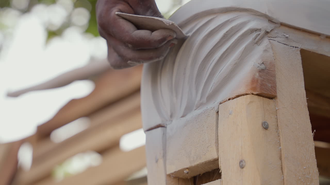 Primo piano di mani nere che modellano una mensola decorativa all'aperto, falegname su commissione che esegue la finitura del legno, movimento costante di levigatura contro il supporto in legno, bokeh illuminato dal sole, progetto del cliente, sensazione tattile.