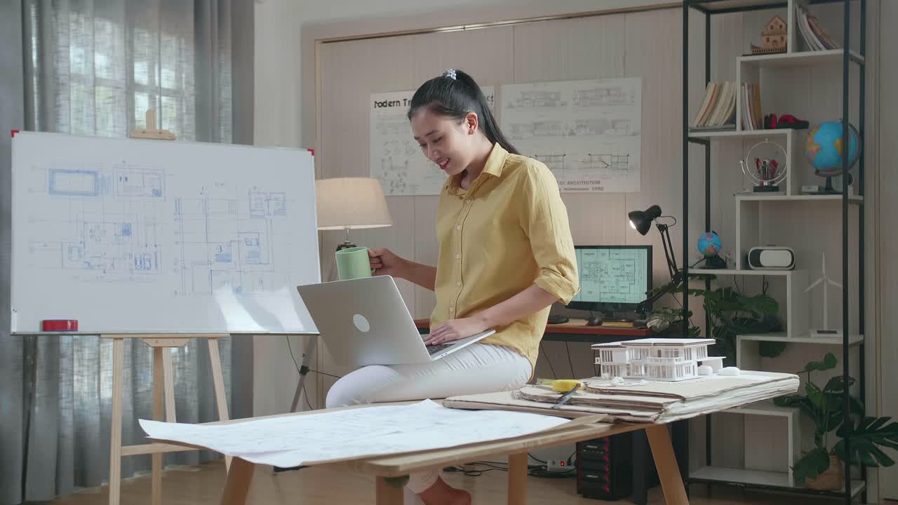 Asian Woman Engineer Sitting On The Table And Drinking Coffee While Using A Laptop To Work At The Office