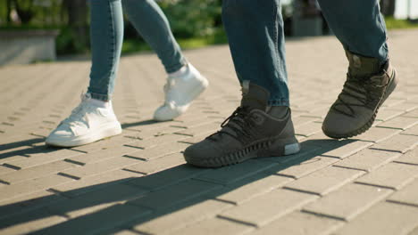 side view of legs in motion on an interlocked pathway, individuals wearing sneakers and boots walking closely together, with greenery and urban structures softly blurred in the background