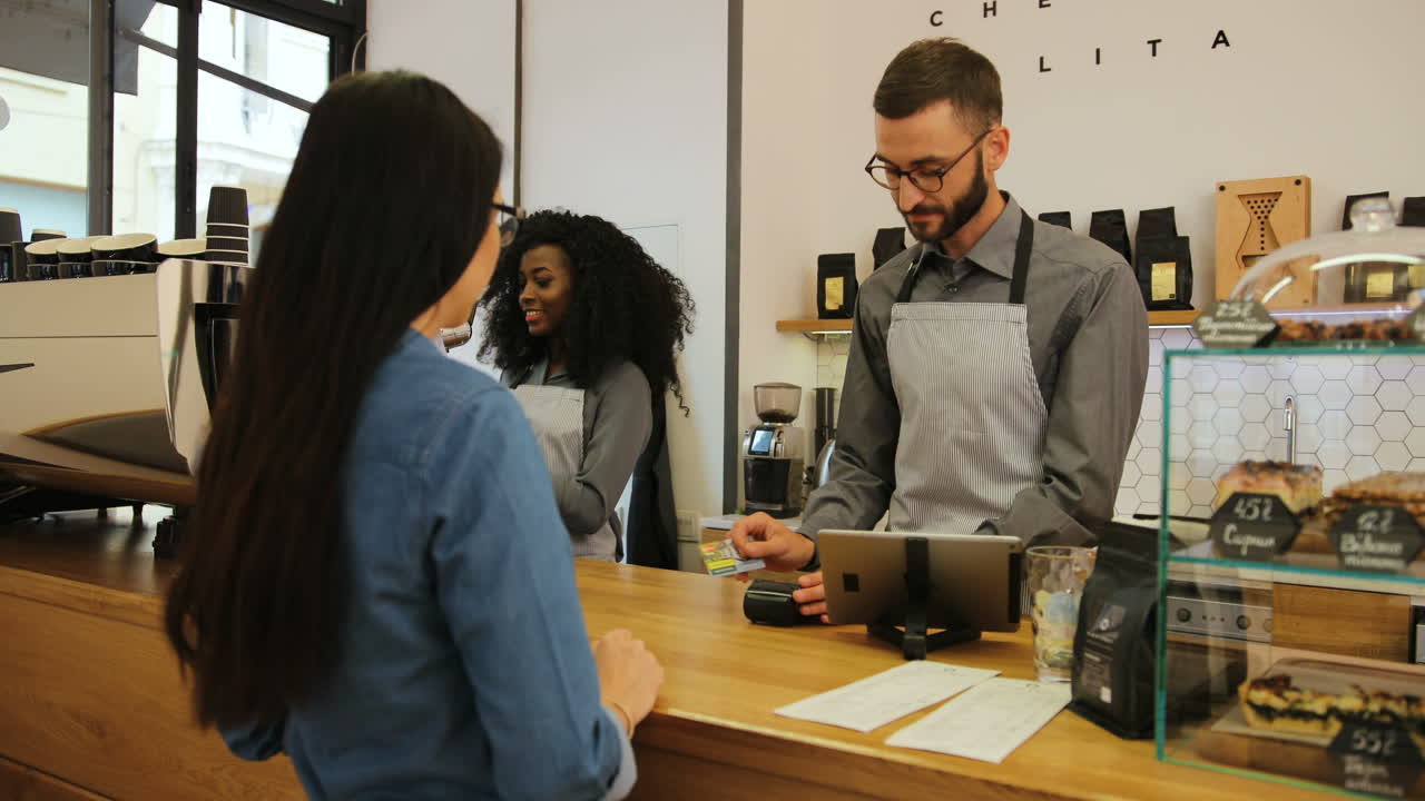 joven caucásica comprando café en una elegante cafetería y pagando con tarjeta de crédito mientras una barista prepara un café