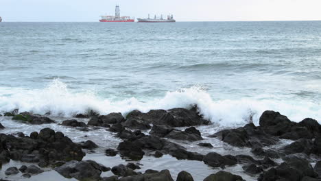 waves crashing into a rocky shore full of large rocks and breaking water leaping to the sides with a pair of boats moored on the horizon at sea 4k slow motion capture at 60fps