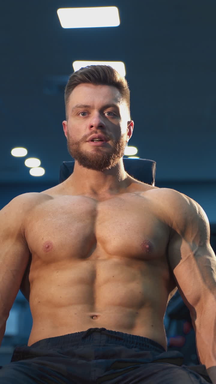 Portrait of a handsome sportsman training in gym. Young bearded sportive guy without shirt sitting in chair and pumping his arm muscles with heavy dumbbells. Vertical video